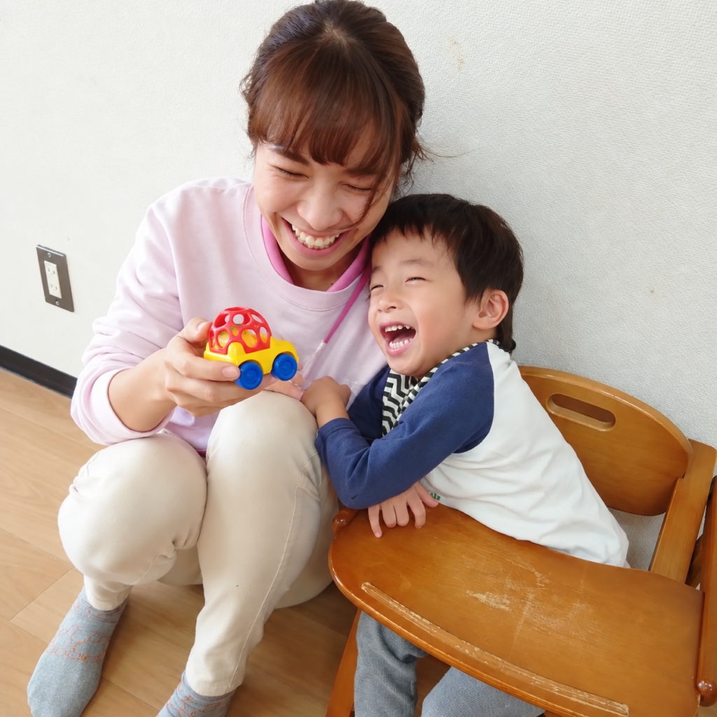 おもちゃの車で遊ぶ幼い子どもと、笑顔で関わるスタッフ